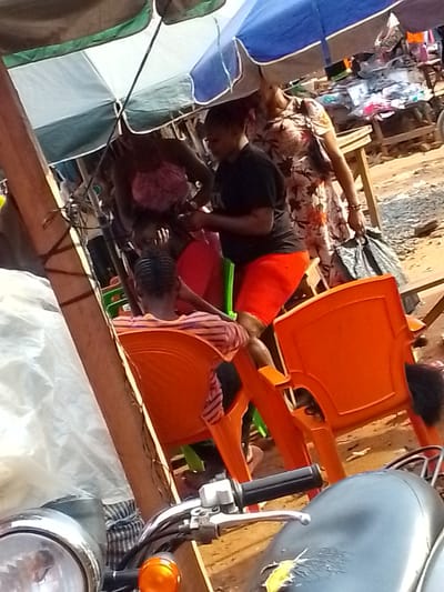The scene is a marketplace setting in Orlu, Nigeria. People are present, with one woman in a floral dress holding a black plastic bag. Another woman is wearing a black shirt and red shorts. There are orange plastic chairs. The area is covered by umbrellas, and merchandise is displayed on tables. A motorcycle is in the foreground, with its handlebars and headlight visible. The ground appears to be dirt.