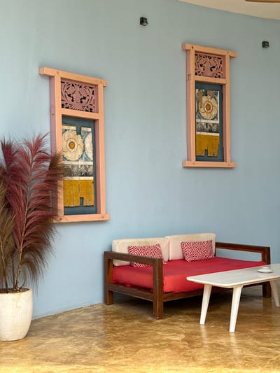 A room with pale blue walls. Two framed wall hangings feature ornate carvings and floral tilework. A wooden-framed daybed with a red cushion and geometric-patterned pillows sits adjacent to a small white table. A tall, feathery plant in a white pot stands near the base of the lower wall hanging. The floor transitions from blue near the wall to beige/yellow. The image is rotated.