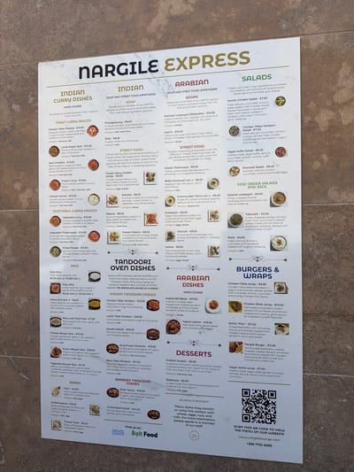 This is a photograph of a menu from Nargile Express, an Indian and Arabian restaurant located in Ta' Xbiex, Malta. The menu is presented in a single, large, landscape-oriented sheet. The top of the menu features the restaurant's name "Nargile Express" in large, stylized lettering. The menu is divided into several sections: "Indian Curry Dishes," "Indian Soup and Street Food Appetisers," "Arabian Dishes," "Salads," "Tandoori Oven Dishes," "Desserts," "Side Order Salads and Rice," and "Burgers & Wraps." Each section lists various dishes with their descriptions and prices in Euros. There are small illustrative images accompanying some of the dishes. Allergy information is provided for some items, and a general allergy statement is included at the bottom of the menu. The menu also displays a QR code for the restaurant's website and contact information. The background of the image is a textured surface, likely a table or countertop, with a subtle pattern. The lighting is bright and even, suggesting an indoor setting. There are no people or specific actions depicted in the image; it is solely a visual representation of the restaurant's menu.