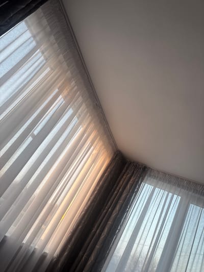 An interior scene in the city of Tsenovo, Bulgaria, captured from a low, upward-angled perspective, shows a corner of a room dominated by windows and curtains. The upper right quadrant features a plain white ceiling. A white curtain rod runs horizontally across the top, from which two layers of curtains are suspended. A sheer, translucent white or off-white curtain extends across the upper left and lower central portions of the image, allowing natural light to filter through. Direct sunlight, appearing with a warm, golden hue, strongly illuminates the lower left section of these sheer curtains, creating vertical patterns of light and shadow. Behind the sheer curtains, and partially visible on the far left and prominent on the right, are darker, opaque curtains, possibly grey or charcoal, exhibiting a subtle geometric pattern. The portion of the room visible through the sheer curtains on the right exhibits a cooler, bluish light. No individuals or specific actions are depicted.