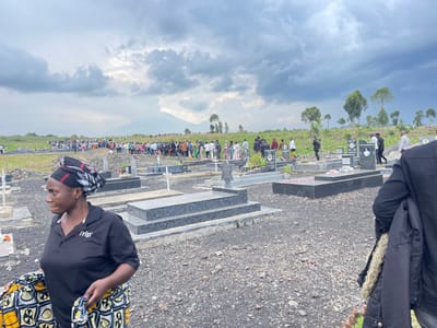 The scene takes place in a cemetery in Nzulo, Democratic Republic of Congo (latitude: -1.601, longitude: 29.123). A large group of people are gathered, walking in a line towards a grave site.  The people are dressed in various attire, suggesting a funeral ceremony. In the foreground, a woman wearing a black shirt with a white "nip" logo and a black and white patterned head scarf stands near the edge of the frame. She is holding a black and yellow patterned cloth in her hands. A man wearing a dark jacket is standing near the far right side of the image, holding a jacket and looking at the ground. The background shows a dirt path leading up to the hill in the distance. There are rows of graves with flowers on them, and a few crosses standing upright. The sky is overcast, with dark clouds hanging in the air.  The environment is quiet and somber, suggesting a solemn occasion.