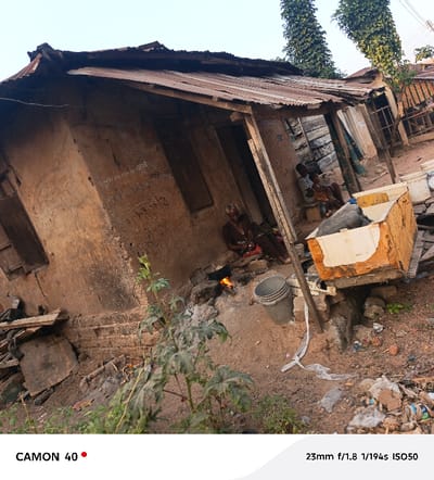 An elderly woman cooks over an open fire outside a dilapidated mud house with a rusty metal roof. Nearby, a bucket sits on the ground. In the background, two people are seated on a bench near a wooden structure. The foreground is cluttered with debris and a plant. A partially painted wooden box is positioned near the house. The image appears to have been taken at an angle, lending a slightly skewed perspective.