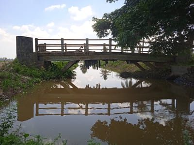 A concrete pedestrian bridge spans a brown, murky canal. The bridge has a weathered appearance, with wooden railings and some debris hanging from its underside. The canal reflects the bridge, the sky, and surrounding trees. The banks of the canal are overgrown with grass and vegetation. A large tree with dense foliage is situated to the right of the bridge. The sky is partly cloudy with a bright, overcast sun. The scene depicts a rural or semi-rural environment in Meunasahblang, Indonesia.