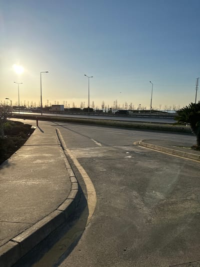 The scene depicts an outdoor roadside environment in Zeytinburnu, Turkey, under a clear sky with the sun low on the horizon, casting long shadows from the upper left. In the foreground, a concrete sidewalk curves to the right, bordered by a dark grey curb. Adjacent to the curb on the asphalt road surface is a yellow painted line. A white arrow is painted on the asphalt, indicating a right turn toward the background. A red fire hydrant is visible within a landscaped area of dark foliage to the left of the sidewalk.

In the midground, a multi-lane highway or major road extends horizontally across the frame. Multiple tall, dark-colored streetlights are positioned along this road. A barrier or guardrail separates the highway from a strip of land beyond it, which is sparsely populated with mostly bare trees. Two vehicles are partially visible on the far side of the highway: a white box truck toward the center-left and a dark-colored passenger vehicle (possibly an SUV or van) further to the right.

The background reveals a body of water, consistent with the Marmara Sea, extending to the horizon. Distant landmass or structures are faintly discernible across the water. No individuals or specific actions are observed in the scene.