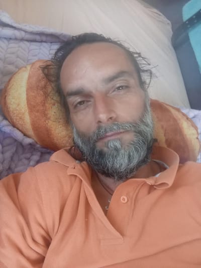 A man with a grey beard and dark hair is lying on a pillow that resembles a loaf of bread. He is wearing an orange polo shirt. The background shows a light-colored blanket or sheet and a teal object. The man is looking directly at the camera. The setting appears to be indoors.