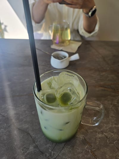 A close-up shot features a clear glass mug filled with an iced matcha latte, complete with ice cubes and a black straw, resting on a dark, textured table. In the background, a person wearing a white shirt is visible, holding a smartphone and a silver smartwatch on their left wrist. A second glass, containing a pale yellow liquid, sits further back on the table, near a small white creamer. The lighting suggests an indoor setting, possibly a cafe in Gumi-si, South Korea.