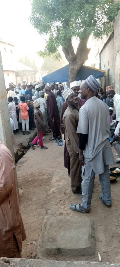 The image depicts an outdoor gathering of approximately 30-50 individuals, predominantly adult males and several boys, within an unpaved thoroughfare in Kabuga, Nigeria. The scene is illuminated by natural daylight, casting soft shadows.

The foreground features an uneven dirt ground. On the lower left, a portion of a man in a light brown striped tunic is visible. A rectangular, weathered stone block is embedded in the dirt slightly to the right of the center foreground. An open, unlined ditch runs along the left side of the dirt path. In the mid-ground, a young boy in a brown long-sleeved shirt and bright pink trousers walks towards the left.

The central focus includes two adult males facing each other and smiling. The man on the left wears a brown long-sleeved tunic, brown trousers, and a patterned light-colored cap. The man on the right wears a light grey short-sleeved tunic over a black long-sleeved shirt, grey trousers, and a blue patterned cap. To their right, several pairs of sandals and slippers, including blue and black ones, are scattered on the ground. Further right, near the edge of the frame, two light-colored bowls are on the ground, one containing yellowish food.

The background shows a dense crowd of people, mostly men, dressed in various traditional tunics and caps (kufis). Colors include white, blue, brown, maroon, and orange. Many are standing and interacting. Behind the crowd, a large tree with green foliage stands prominently, its trunk visible above a blue tarp or awning that extends over part of the gathering. Buildings with light-colored walls line both sides of the street; the structure on the left features arched windows, while the building on the right has a simpler, unadorned facade.