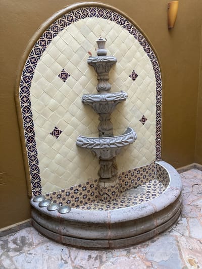 Here is a detailed description of the image:
**Location:** The image depicts a decorative water fountain, likely situated in a building or courtyard setting in Tres de Mayo, Mexico.
**Environment:** The fountain is built into a wall with a painted tan or ochre surface. Adjacent to the fountain, the wall transitions to a rough, stone-like texture with visible weathering and discoloration.
**Objects:**
* **Fountain basin:** A semi-circular basin is the focal point, lined with cream-colored square tiles adorned with small, dark floral patterns. A border of similar, more intricately patterned tiles surrounds the main tiled area.
* **Sculptural elements:** Three decorative, grey stone pillars or supports are centrally positioned within the basin area. They feature ornate carvings and a weathered appearance, suggesting age.
* **Water spout:** A small water spout is visible at the top of the fountain.
* **Arched frame:** The fountain is framed by a multi-layered stone archway, which appears to be made of concrete. A series of four metallic-looking, disc-shaped objects are affixed to the side of the frame.
**Subjects:**
The subject is the architectural design of the fountain.
**Actions/Interactions:** None are explicitly visible. It is implicit that the fountain's intended function is to circulate water.
**People:**
There are no people present in the image.