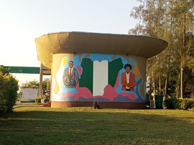 The image depicts a cylindrical, tan-colored structure with a wide, overhanging roof, located outdoors in Tawpo, Nigeria. The front of the structure features a mural with two portraits flanking a vertical green, white, and green stripe resembling the Nigerian flag. To the left of the flag is a man wearing a suit and tie, and to the right is a woman in a blazer. The portraits and flag are surrounded by a colorful, abstract design.
The structure is situated on a well-maintained lawn. To the left is a covered walkway and a fenced area with a sign. To the right, trees and shrubs are present, along with a green trash can and an air conditioning unit. A tree stump is visible in front of the structure.