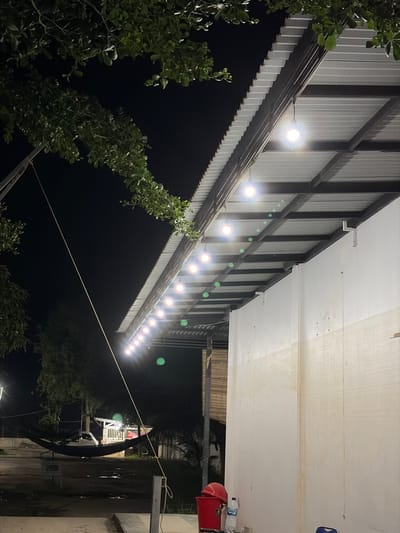 This photograph depicts an outdoor area at night, possibly a restaurant or a gathering space, located in Tanahterban, Indonesia. The scene is illuminated by a string of hanging lightbulbs suspended from a corrugated metal roof. The roof extends over a light-colored wall, which shows some signs of dirt or discoloration. To the left of the frame, green foliage from trees drapes over the scene, with a dark night sky visible behind it. In the background, a black hammock is strung between two points, with a white vehicle parked nearby. A red bucket and a plastic water bottle are visible at the base of the wall in the foreground. The ground appears wet or reflective, suggesting recent rain or a damp environment. The overall impression is one of a simple, rustic outdoor setting in the evening.