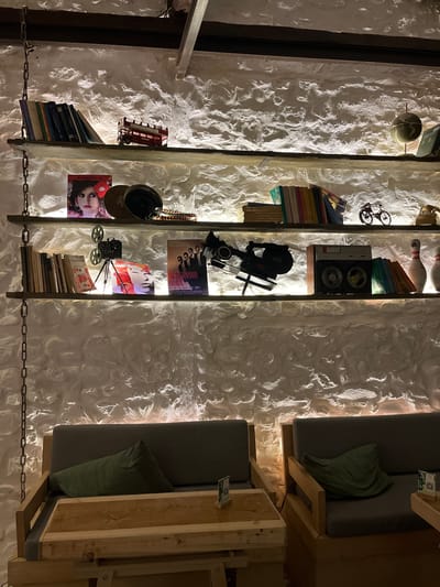 The image shows an interior wall in Xanthi, Greece. Three vertically stacked shelves are displayed on the left, built into a white textured wall. The shelves are filled with various objects, including books, film equipment, model vehicles, and decorative items. To the right of the shelves are two built-in seating areas with gray cushions and green pillows. Each seating area has a small wooden table with a card. A chain is visible near the bottom of the image, running horizontally. The lighting suggests an indoor setting with artificial light sources.