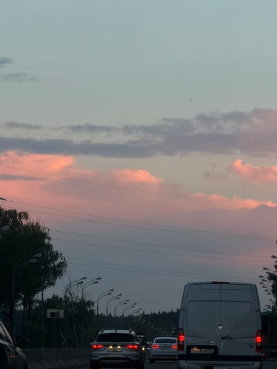 The image depicts a highway scene at dusk, with a traffic jam. The sky is a gradient of pale blue and soft pink, with wispy clouds illuminated by the setting sun. Numerous streetlights line the right side of the road, and power lines crisscross the sky. Several cars are visible on the highway, including a silver SUV in the foreground with its brake lights on, and a white van behind it. The SUV has the license plate M608VO 777. The white van has the license plate C223YA 797. Trees and foliage line the left side of the highway. The location is identified as Krasnaya Pakhra, Russia.