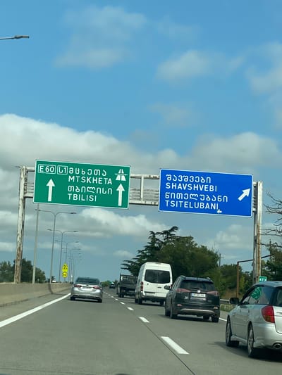 The image shows a highway scene in Kvemo Shavshvebi, Georgia, with directional signs indicating routes to Mtskheta, Tbilisi, and Tsitelubani. Two large green and blue overhead signs are prominently displayed. The green sign, on the left, displays "E60 L1" followed by Georgian script and "MTSKHETA TBILISI" with upward arrows. The blue sign, on the right, shows Georgian script and "SHAVSHVEBI TSITELUBANI" with a rightward arrow. Several cars are on the highway, including a silver sedan, a black SUV, a white van, and a truck. The sky is partly cloudy with a bright blue expanse. A yellow speed limit sign with "60" is visible on a pole to the left. The environment appears to be a typical highway with concrete barriers and streetlights.