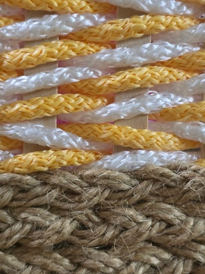 The image shows a close-up view of a woven material composed of thick ropes. The top portion of the weave features alternating bands of yellow and white braided ropes, interspersed with thin wooden slats. Below this, the weave transitions to a thicker, natural fiber rope in a brown, earthy tone, also appearing to be braided. The texture is rough and fibrous. The scene is an indoor shot focusing solely on the textile. There are no people, city locations, or any environmental context visible.