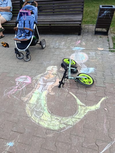 A daytime, eye-level, full shot captures a paved area with chalk drawings on the ground. In the foreground, a green balance bike lies on its side, with its wheels pointed upwards and handlebars to the left. Surrounding the bike and extending to the left are chalk drawings depicting a mermaid, a jellyfish, and other abstract shapes in various colors including pink, green, yellow, white, and blue. A blue stroller is positioned in the mid-ground to the left, with its wheels visible. Behind the stroller, a dark wooden park bench runs horizontally across the frame. To the far left, a person's legs and torso are visible, seated on the bench, wearing a light blue denim outfit. In the background, to the right, a dark-colored trash can stands upright. The ground is paved with interlocking reddish-brown bricks, and green grass is visible beyond the bench and trash can. The scene appears to be outdoors, possibly in a park or public space within the city of Gubkin, Russia.