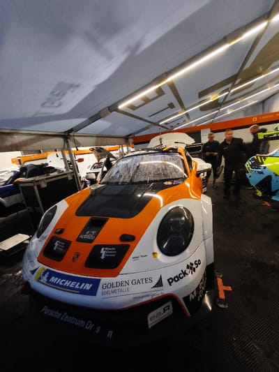 A forensic examination of the image reveals a detailed view inside a temporary pit garage or service tent, located in Flatschach, Austria.

The primary subject is a Porsche 911 GT3 race car, prominent in the foreground and oriented towards the viewer. Its livery is predominantly white and orange, with a black hood and roof section. Numerous sponsorship decals are visible, including "MICHELIN," "GOLDEN GATES EDELMETALLE," "Pack So," "Porsche Zentrum Dr. S N," "ea.R," "Moschs Elektro Service," "HERRANDO," and "ADAC Team." The car's windshield and front bodywork are covered in small water droplets or condensation. Faintly visible on the right headlight cover is the number "17." The vehicle rests on a dark, possibly checkered, floor surface.

In the mid-ground, three adult males are standing behind the main race car. They appear to be team personnel, dressed in dark attire, and are observing the scene. One individual on the left is holding what appears to be a tablet or clipboard. To the left of the main car, a large, dark equipment case is present. Further in the background, other race cars or car components are partially visible, including a white vehicle with its hood open and a distinct blue and green livery car on the right side of the frame.

The environment is illuminated by linear LED light strips affixed to the underside of a light-colored, possibly white, canvas tent ceiling. Support beams for the tent structure are also visible. Shadows of lettering, including "CASTROL," are reflected on the tent ceiling. The background walls or partitions are predominantly white, indicating a modular or temporary structure.