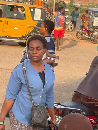 This image captures a bustling street scene, likely in an urban or semi-urban environment, under warm, golden hour lighting, suggesting either early morning or late afternoon. The sun casts distinct shadows on the ground.
In the immediate foreground, positioned to the right, a woman with short, dark, curly hair looks directly towards the viewer. She is wearing a light blue long-sleeved collared shirt over a black undergarment (possibly a bra or camisole) and a patterned skirt with brown, white, and blue hues. A brown shoulder bag with a distinct monogram pattern, resembling a Louis Vuitton design, hangs across her body. On her left wrist, she wears a black band or bracelet with white text that appears to read "UNITS". Her expression is neutral.
Behind and slightly to the left of her, another woman with dark hair is partially visible, wearing a dark blue garment with white horizontal stripes. She appears to be carrying a small child, possibly a baby or toddler, whose legs in a matching striped outfit are visible emerging from behind her back.
Further into the mid-ground, to the left, a man is standing next to a yellow three-wheeled commercial vehicle, often referred to as a 'Keke Napep' or auto-rickshaw. He is wearing a colorful tie-dye t-shirt (red, blue, and yellow) and red shorts. Near him are several tires. Inside the yellow vehicle, through the window, at least two people are visible, one smiling broadly.
Behind the yellow vehicle and the man in the tie-dye shirt, another man is seen walking, carrying a large, bundled item on his head. He is wearing a light blue or greenish shirt and dark pants.
In the background, the street is populated with several people and vehicles. A red and white motorcycle is parked or being ridden by a person in a patterned dress. A white van is also visible further back, along with other motorcycles. On the far right, a partial view of a person in a dark top and a motorcycle seat is visible, along with a rounded brown object.
The overall impression is one of daily life unfolding on a busy thoroughfare, with people moving, transporting goods, and interacting.