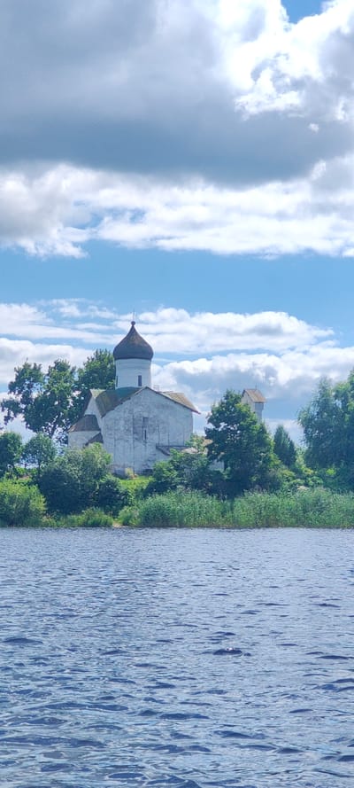 A white church with a dark dome sits on a grassy knoll overlooking a blue body of water. The church has a pitched roof and a simple facade. Trees and reeds surround the church. The sky is blue with scattered white clouds. The water in the foreground is choppy. The image is taken from a low angle, possibly from a boat on the water. The location is the village of Shchiglitsy, Russia.