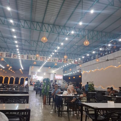 This media file depicts the interior of a dimly lit restaurant or cafe in Namplohbaro, Indonesia. The scene is captured from a high angle, looking up towards the corrugated metal ceiling supported by a green metal frame. Numerous woven pendant lamps and string lights illuminate the space, creating a warm and inviting ambiance.
Several people are seated at tables, engaged in dining or conversation. In the foreground, a woman with dark hair is looking down at her phone. Further back, a man in a blue checkered shirt stands, and a group of people are seated at tables. In another area, a man in a blue t-shirt is holding his phone. People are dressed casually, suggesting a relaxed atmosphere.
The restaurant features rows of tables with black chairs. Decorative elements include large potted plants and framed pictures on the walls displaying what appear to be food and beverages. The overall impression is that of a busy, casual dining establishment, likely operating in the evening or at night, given the lighting. There is some visible text on banners or posters on the wall, but it is not clearly legible. The ambiance suggests a pleasant social gathering or meal.