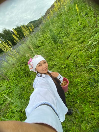 A selfie taken by a woman in a white shirt and a floral headscarf, holding a pink bucket full of berries, is depicted in a grassy field with yellow and purple wildflowers. The woman is smiling at the camera. The location is described as Maloarslangulovo, Russia. The background shows a hill covered in green foliage and a cloudy sky.