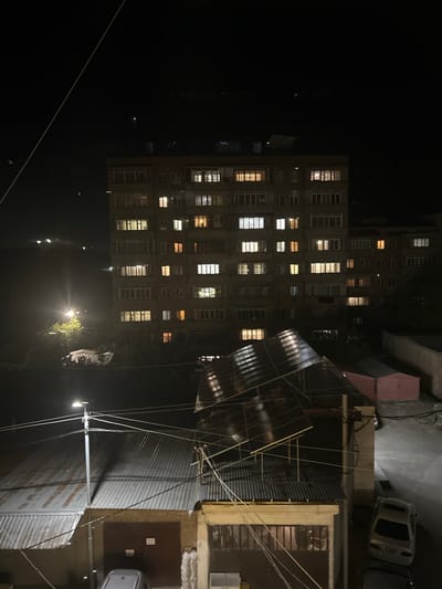 The image displays a nighttime urban scene in Charentsavan, Armenia, captured from an elevated perspective looking down and across.
In the foreground, a large, sloped structure with dark, possibly translucent or solar panels extends from the lower-right to the center. Beneath it, a building with a prominent brown garage door is visible, flanked by corrugated metal roofs. A grey utility pole equipped with a square LED streetlight stands in the lower-left quadrant, casting a bright illumination onto adjacent structures and a portion of a corrugated roof. Multiple utility wires traverse the frame, with one light-colored, thick wire running diagonally from the top-left towards the lower-right.
The mid-ground is dominated by a multi-story residential building, approximately 9 to 10 floors high, with a grey-brown facade. Numerous windows are illuminated with varying intensities and hues of warm and cool light, suggesting interior habitation. To the left of this main building, a bright light source, likely another streetlight, illuminates a small, leafy tree and parts of another building. On the far right, a small, reddish-pink shed is situated near a paved surface.
Three vehicles are visible in the lower section of the image: a light-colored sedan parked facing away from the viewer on the right, a partially obscured dark vehicle behind it, and another partially visible dark vehicle near the bottom-center below the garage door.
The background consists of dark, indistinct landforms, likely hills or mountains, on the left, marked by a few distant, faint lights. The sky is dark, devoid of stars. No individuals are visible, and no specific actions or interactions are depicted within the scene, which is entirely illuminated by artificial light sources.