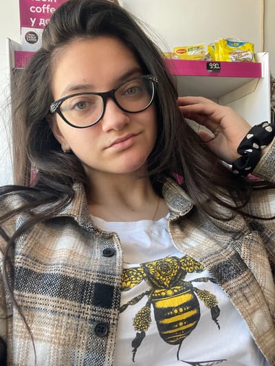 A woman with long, dark brown hair, wearing black-framed glasses and a plaid overshirt over a white t-shirt with a bee design, poses indoors. She is looking at the camera. She has a hair tie with white polka dots on her hand. The background shows a product box with a price of 9.90 and a coffee product box, with "coffee" visible. The exact location within Kraishte, Bulgaria cannot be determined from the image.