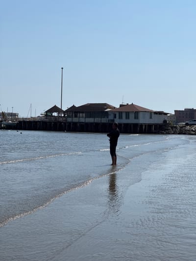The photograph, taken in Anzio, Italy, depicts a person standing ankle-deep in water along the beach, adjacent to a structure resembling a pier or boardwalk extending over the sea. The individual, silhouetted in dark clothing, is positioned parallel to the pier. To the left of the subject, a series of buildings are visible along the shoreline. A tall, thin pole stands near the pier. The environment suggests a coastal setting.
