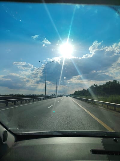 The image captures a view from inside a vehicle looking forward onto a highway in the city of Prikubanskiy, Russia. The sun is bright and high in a clear blue sky, with scattered white clouds. Sunbeams radiate outwards, creating a lens flare effect. The highway has multiple lanes, separated by white lines, and a yellow line on the right side. Guardrails run along the sides of the road, and streetlights are visible at intervals. To the right of the highway, there is a dense line of trees. In the distance, there are faint signs of urban infrastructure and some vehicles on the road. The interior of the vehicle, including the dashboard and a speaker, is visible in the foreground, obscuring the bottom portion of the scene.