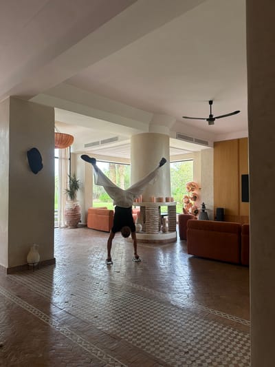 In a bright, spacious living area with high ceilings and large windows overlooking a lush green landscape, a person is performing a handstand. The individual, wearing a black t-shirt and loose white pants, is balanced on their hands with their legs spread wide in a V-shape. The floor is tiled with a mosaic pattern of terracotta and white ceramic. Orange sofas are positioned in the background, and a circular, multi-tiered fireplace with candles sits in the center of the room. A large, textured pot with a plant stands near a pillar on the left. A ceiling fan is visible overhead. The location appears to be Al Ouidane, Morocco, suggested by the architectural style and the surrounding vegetation.