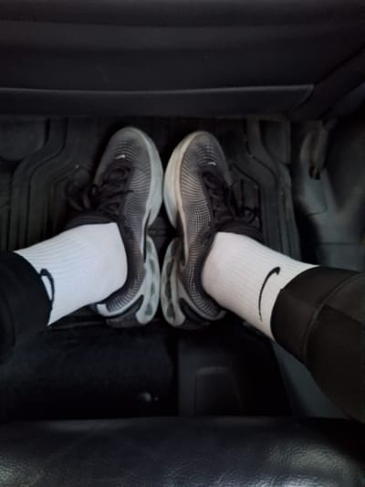 This image depicts a pair of human feet resting within the passenger compartment of a vehicle in the city of Topoli, Bulgaria. The feet are clad in white crew-length socks, each displaying a black Nike swoosh logo. Both feet wear dark grey or black athletic shoes featuring black laces and distinctively segmented, light-colored soles, particularly visible on the outer and rear aspects. Dark apparel, likely trousers or leggings, is visible above the socks, covering the lower legs. The vehicle's floor is covered by dark, textured rubber or plastic floor mats with raised ridges. The background consists of dark, likely upholstered, surfaces consistent with an automotive interior. The perspective is downward-looking, capturing the footwell area of the vehicle.