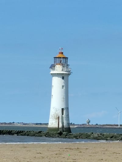 The image shows a lighthouse on a beach in Wallasey, United Kingdom. The lighthouse is white with a red and yellow top and has a ladder on the side. There is a breakwater made of rocks in front of the lighthouse, and the sea is visible beyond that. In the background, there are some buildings and a wind turbine. The sky is blue and clear.