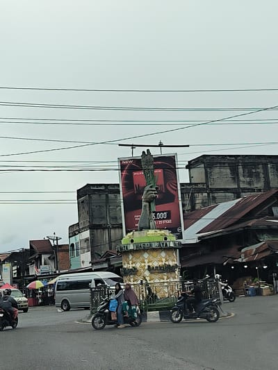 The image captures a street scene in Simpangpeut, Indonesia, on an overcast day. In the foreground, a paved road shows multiple lanes of traffic, with a roundabout area in the center. Several motorcycles are present, with one carrying two women wearing headscarves. Another motorcycle is ridden by a man in black. Further down the road, a white van is parked.

Dominating the central background is a large billboard for Marlboro cigarettes, featuring a hand holding what appears to be a stalk of tobacco. In front of the billboard is a monument, decorated with intricate patterns and topped with a green, crown-like structure. The monument is illuminated by yellow lights and has a metal railing around its base.

Behind the monument and billboard, several buildings are visible. Some appear to be old and weathered, with peeling paint and concrete facades. Others have corrugated metal roofs, suggesting a bustling marketplace or commercial area. The sky is a uniform gray, and numerous power lines crisscross the upper portion of the image. Various signs, including one with the letters "OSI", are also discernible on the buildings. The overall impression is one of an active urban environment in a tropical region.