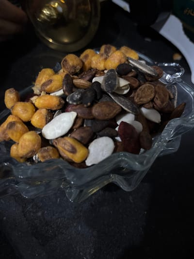 A clear, irregularly shaped glass bowl, with a wavy top rim, is centrally positioned on a dark, textured surface, likely a tabletop. The bowl contains a mixed assortment of roasted snacks. Visible items include golden-yellow roasted corn kernels or chickpeas, dark brown and black watermelon or pumpkin seeds, striped gray and white unshelled sunflower seeds, and white shelled sunflower or pumpkin seeds. Some darker, possibly red-hued seeds or nuts are also interspersed within the mixture. In the blurred background to the upper left, a golden metallic object, possibly a cup or a decorative item, is partially visible, with a faint suggestion of a human hand near it. To the upper right background, a blurred dark green object with white Arabic script is visible. The scene is located in Mu'adammiyat ash-Sham, Syria.