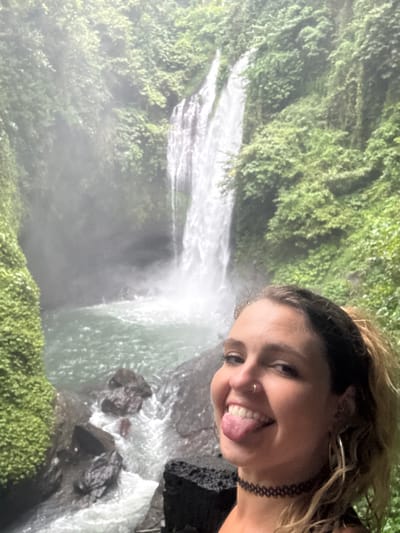 A young woman smiles with her tongue out, posing for a selfie against a backdrop of a vibrant waterfall. The cascading water is partially obscured by mist, surrounded by lush green foliage. The woman wears a choker and an earring, and her hair is tied up. The photo has been rotated, placing the waterfall on its side.