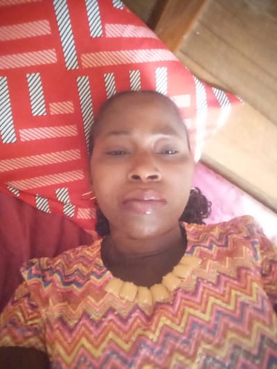 The image displays a close-up, eye-level perspective of an adult female subject positioned indoors, appearing to be lying down. The female has dark-toned skin, dark eyes, and dark, curly hair visible on the right side of her head. Her lips are slightly parted and appear glossy. She wears small, gold-colored hoop earrings. Her garment is a patterned top featuring a multicolor zigzag (chevron) design in hues of red, orange, yellow, blue, and white. The neckline is adorned with distinct, light yellow/tan, rounded rectangular elements. A dark-colored layer is visible beneath the patterned top's collar. Behind her head is a red pillowcase with a repeating geometric pattern of white and black striped rectangles and diagonal lines. A wooden surface is visible in the upper right background, and a solid dark red fabric object is present in the lower left. The scene is located in Unguwan Rimi, Nigeria.