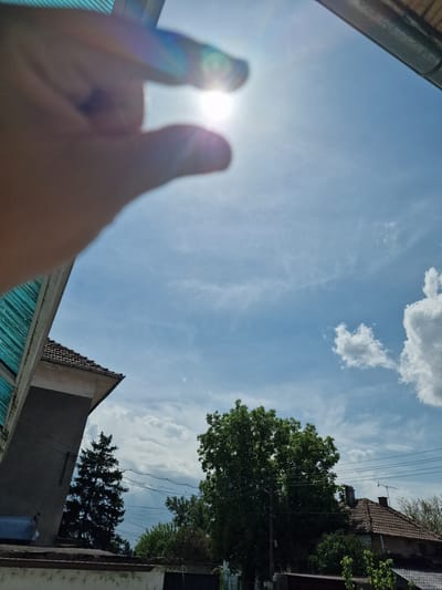 The image is a low-angle shot of a sunny sky with scattered clouds, taken from what appears to be an enclosed or semi-enclosed area. In the foreground, a blurred hand is positioned to frame the sun, creating a heart shape with the fingers. The surrounding environment includes rooftops and trees, typical of a residential or suburban area. The architectural styles visible suggest a location within a Central European or Balkan context. Given the specified location of Byala Slatina, Bulgaria, the scene is likely within a residential area of that city. Overhead power lines run parallel to the roofline in the distance. The angle implies the photographer is either looking up from a low vantage point, or the camera is angled upwards.