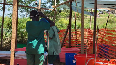This outdoor image depicts two individuals, likely healthcare workers, within a makeshift shelter in a lush green environment. The shelter is constructed from wooden posts and a light blue tarpaulin roof. The scene is set under a bright, sunny sky, with dense foliage and trees forming the background. In the distance, through the trees and an orange plastic mesh fence, several simple buildings or huts are visible.

The person in the foreground, seen from behind, wears green scrubs and is adjusting something on their head, possibly a mask or headscarf. Another individual, also in green scrubs, is partially visible behind them, bending over, engaged in an unseen task. On the ground, numerous plastic tubs and buckets in various colors, including pink, blue, red, and orange, are arranged. Many appear to contain water, suggesting activities related to washing, sanitation, or water storage. Orange plastic safety fencing forms a barrier around part of the area, and rebar rods are also present.

The setting indicates a temporary or semi-permanent facility, likely for health services or hygiene, within a rural or camp environment. The location context is Tchere, Democratic Republic of the Congo. The phrase "Joseph Kisuki" is visible as a watermark in the bottom right corner of the image.