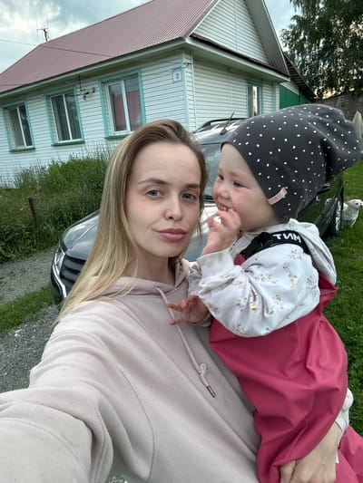 A woman and a baby are in the foreground of this selfie. The woman has long blonde hair and is wearing a light pink hoodie. The baby is wearing a dark gray polka-dot hat and a white hooded sweatshirt with a floral pattern and a pink dress. The baby is looking towards the woman, and the woman is looking at the camera. The background shows a house with white siding and green trim, a car, and grass. The scene appears to be outdoors in a rural or suburban setting. The lighting suggests it is daytime. There is text visible on the baby's sweatshirt that appears to be Russian letters and reads "ТИМ". The location is indicated as Азинский, Russia.