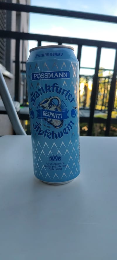 The image shows a blue can of "Possmann Frankfurter Apfelwein Gespritzt" (sparkling apple wine) lying horizontally on a light-colored surface. The can features a traditional German ceramic jug design and text indicating it's a prized apple wine from Germany. The background includes a dark metal structure and blurry green foliage, suggesting an outdoor setting on a balcony or patio. The scene appears to be an ordinary, unidentifiable location in Germany, but not specifically Rüsselsheim am Main.