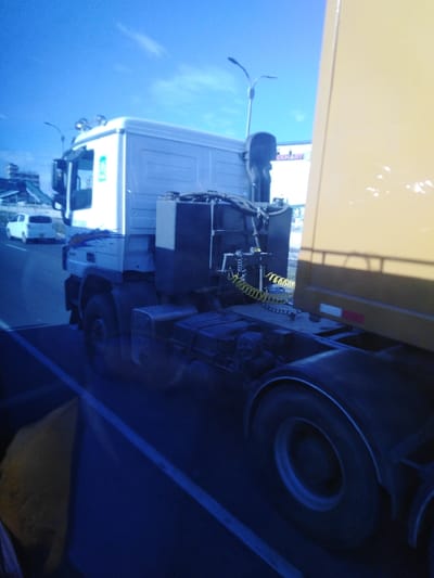 A white cab-over truck with a yellow trailer is on a road, a white car is visible ahead on the left. Streetlights are visible and what looks like a billboard or sign is also in view. The image is taken through a bluish tint, possibly a window. The truck's black equipment box behind the cab is prominent, as is the yellow coiled trailer cord. A portion of what could be a seat is in the lower left corner.