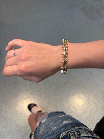 In this close-up, overhead shot, the focus is on a person's left arm and hand, adorned with jewelry. A thin, gold-colored bracelet with a repeated star-like or asterisk-like pattern encircles the wrist. On the ring finger, a delicate, thin band with a textured or bejeweled edge is visible. The skin tone suggests fair complexion. The background is a neutral, mottled grey surface, possibly a floor or countertop, with a slight sheen. To the right of the arm, a portion of distressed denim jeans is visible, suggesting casual attire. The lighting appears to be natural or well-lit indoor lighting, creating soft shadows and highlighting the metallic sheen of the jewelry. The overall impression is one of a stylish detail being showcased, with no particular activity or event strongly suggested, other than perhaps posing or displaying the accessories. No text is visible in the image.