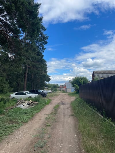 The image depicts a rural or semi-rural unpaved dirt road in Vasilevka, Russia, under a clear blue sky with scattered white cumulus clouds, indicating daytime. The road, showing tire tracks and small pebbles, extends from the bottom center into the background.

On the left side of the road, a dense line of tall coniferous trees (pines) dominates the upper and mid-ground, casting shadows. Beneath these trees, three vehicles are parked sequentially: a white sedan, a black SUV, and a lighter-colored sedan further back. The white sedan, closest to the foreground, displays front-end damage, including a dented bumper/fender. Adjacent to the white sedan and partially obscured by tall grass and weeds, a pile of discarded wooden planks and debris rests on a white sheet or tarp.

On the right side of the road, a strip of tall green grass and weeds borders the immediate foreground. Beyond this vegetation, a tall, dark brown corrugated metal fence runs parallel to the road, extending into the background. Behind the fence, a building with a dark, corrugated roof and light-colored walls is visible. Further in the background, additional utilitarian structures, including concrete block buildings and a red-roofed structure, are discernible. Power lines are faintly visible against the sky in the upper right quadrant. No individuals or animals are present in the scene.