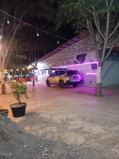 The image depicts an outdoor scene at night, likely a roadside establishment in Cotkumbang, Indonesia, characterized by a dirt and gravel ground surface. A light-colored car is parked on the right side of the frame, facing towards the left. Behind the car is a building with a white slatted facade, illuminated by pink neon lights along its base. A sign with text, partially obscured by a tree, is visible on the building. In the foreground, a potted plant with green foliage sits on the ground to the left of the parked car. The background features trees with visible branches and leaves, strung with decorative lights. The overall ambiance suggests a casual dining or gathering spot.