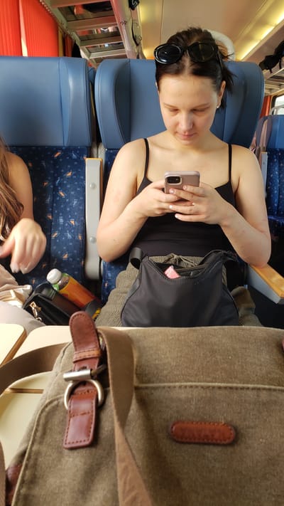 A woman, with dark hair pulled back and sunglasses resting on her head, sits on a train. She is wearing a black tank top and green pants, and is holding a smartphone in her hands, looking down at the screen. Her arms have some red marks on them. A black purse rests on her lap, with a pink item protruding from it. A brown canvas bag with leather accents is in the foreground. Behind her, blue train seats are visible. The scene is lit with artificial light. There is no indication of the specific location in Púchov, Slovakia.