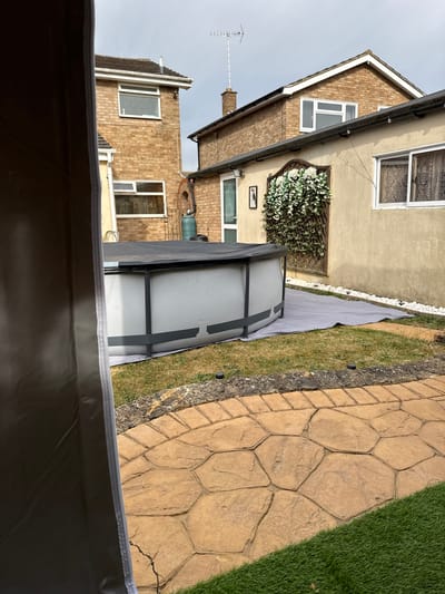 The image shows a section of a backyard in what appears to be Colchester, United Kingdom. A rectangular, above-ground swimming pool with a grey frame and white liner sits on a grey rectangular covering placed on the grass. The pool is positioned close to a house with two sections – one section has beige siding and a window with curtains, with an ornamental flower arrangement beside it, and the other section is brick. A cobblestone path is located next to the grass. Part of a grey covering occupies the foreground, obscuring the lower part of the image. The sky is overcast.