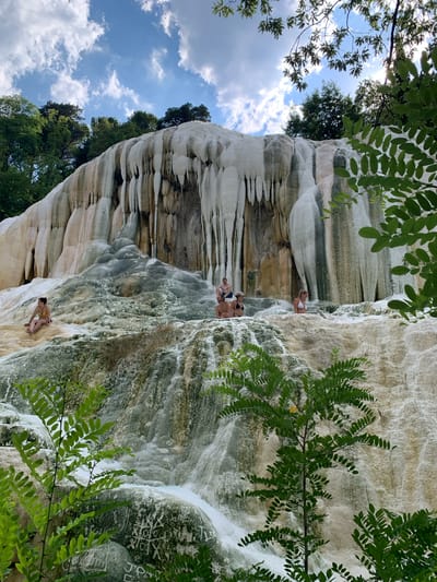 In this vertically oriented photograph, several individuals are positioned on and around a large, mineral-rich formation, likely the White Whale of Bagni San Filippo, Italy. The formation is composed of white and off-white calcium deposits, with streaks of green algae and dark mineral stains, suggesting the presence of flowing thermal water. The texture of the rock is varied, with smooth, cascading areas and rougher, more textured sections.

The subjects, dressed in swimwear, are interacting with the environment. Some are seated or resting on ledges, while others appear to be climbing or exploring the mineral deposits. One person is visible near the top of the formation, leaning against it. Further down, a group of three people are seated together. Another individual is situated lower down, facing away from the camera, with their back to the viewer. The people appear relaxed, suggesting they are enjoying the natural thermal springs.

The setting is outdoors, with lush green foliage framing the upper and right edges of the image, implying a natural, possibly forested, environment. The sky, visible in the upper left portion of the image, is a vibrant blue with scattered white clouds, indicating fair weather and likely daytime. The overall impression is one of a natural wonder being explored and enjoyed by visitors. There is no visible text within the image.