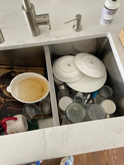 A double stainless steel sink in a kitchen located in Rye, New York at coordinates 40.952, -73.708. The sink is filled with dirty dishes. The left side of the sink has a white bowl filled with brown liquid. The right side of the sink is filled with various dishes, glasses, and other kitchen items. There are 4 white plates stacked on top of each other, a white mug, 3 glass glasses, a blue cleaning brush, and various glass jars. There are also two soap dispensers on the countertop above the sink. The soap dispensers are made of stainless steel. The countertops are white and made of marble. The floor is made of wood. There is a foot in the bottom right corner of the image. There are no people in the image.