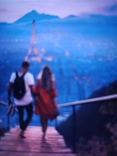 The image is a blurred, low-light shot of two people walking down wooden steps at dusk. The man on the left wears a white t-shirt and a black backpack, while the woman on the right is in a red dress. In the background, the lights of a city twinkle, and a mountain range is visible, with a tower that resembles the Eiffel Tower standing prominently. The scene suggests a romantic or scenic overlook, possibly in Balakliya, Ukraine, given the context. The overall impression is one of a serene and picturesque evening.