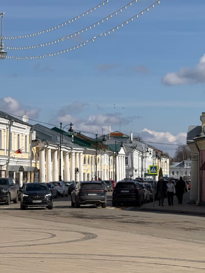 The image depicts a street in Novo-Nikol'skoe, Russia, lined with classic buildings featuring white columns. Cars are parked along the right side of the street, with several dark-colored SUVs visible. People are walking on the sidewalk on the right side of the image. Overhead, strings of decorative lights are strung across the street. The sky is blue with scattered clouds. A crosswalk sign is visible. There are tire marks on the road.