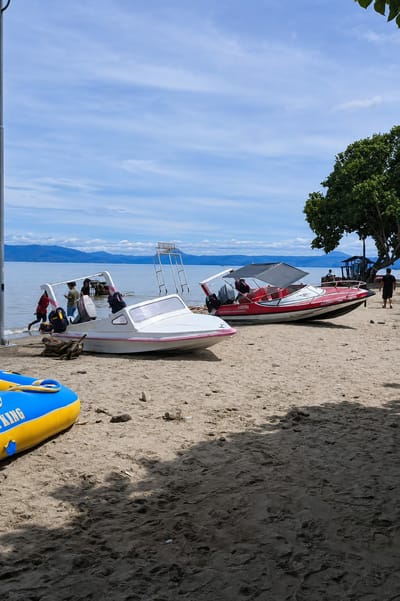 In Situngkir, Indonesia, on a bright, sunny day with scattered clouds, a sandy beach is lined with several small motorboats. In the foreground, a partially visible yellow and blue inflatable raft rests on the sand. Further back, a white motorboat with pink trim is pulled ashore, with several people standing near its stern. To its right, a red and white motorboat with a canopy is also beached, with two individuals visible inside. In the background, the calm, blue water of a large lake stretches to a distant, hazy mountain range. A lifeguard stand, a cluster of trees, and some distant structures are also visible along the shoreline.